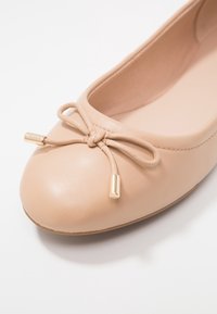 Ballerina in pelle beige con punta arrotondata, con dettaglio a fiocco e accenti in metallo dorato alle estremità del fiocco. Texture liscia.