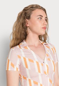 Vestido de mangas curtas em rosa com riscas diagonais brancas e laranjas, com colarinho e botões. Textura suave e canelada. Brincos de argola dourados.