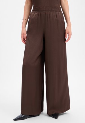 Een persoon draagt losse donkerbruine wijde broek met elastische tailleband en zwarte spitse schoenen, staand met ontspannen handen langs het lichaam.
