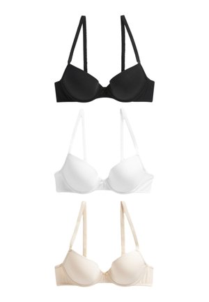 Kvinde med lys hud og langt hår iført en bh i nude-farve, stående mod en ensfarvet baggrund, kigger let til siden.