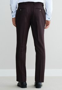 Pantalon pour homme de couleur bordeaux foncé, cintré, avec une texture lisse, deux poches arrière et des détails de couture subtils, accompagné de chaussures noires.