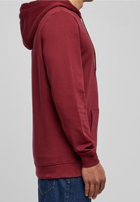 Hombre que lleva una sudadera burdeos con bolsillo frontal y jeans azules, de pie de lado contra un fondo gris liso.