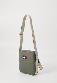 Borsa a tracolla rettangolare verde oliva in tessuto, dotata di chiusura con zip, tracolla regolabile beige e patch con logo sul davanti.