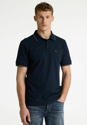 Fiatal férfi tengerészkék galléros pólóban, fehér szegéllyel és kék farmernadrágban, egyik kezével a zsebében áll, egyszínű fehér háttér előtt.