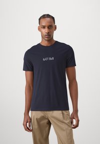 Camiseta de manga corta color azul marino hecha de algodón, con el logo bordado "GUESS" en blanco y azul claro en el pecho.