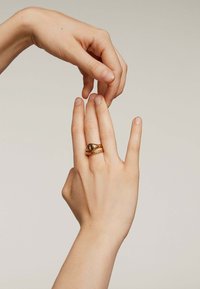 Anillos de oro superpuestos en una mano; uno de los anillos presenta una forma de lágrima con un diseño central. Textura de piel suave y fondo neutro.