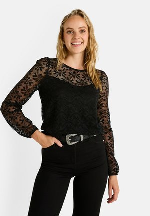 Blouse en dentelle noire avec des manches longues et bouffantes, présentant un motif en vagues. Portée sur une tenue noire simple, assortie d'une ceinture noire texturée.