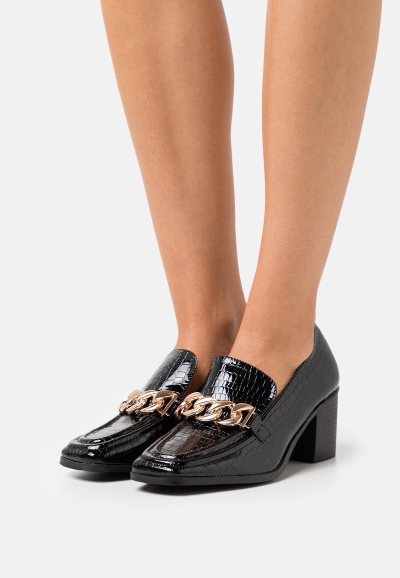 Mocassins à talon bloc noirs avec embossage crocodile et chaîne dorée, portés sur des jambes nues sur un fond clair uni.