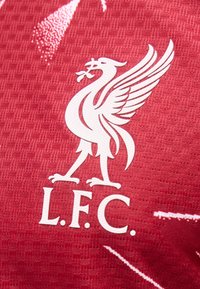 Λευκό λογότυπο Liver bird με το κείμενο "L.F.C." σε υφασμάτινη επιφάνεια κόκκινου χρώματος με υφή, μέρος αθλητικής φανέλας.