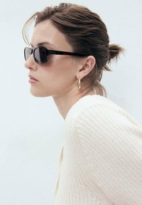 Black rectangular sunglasses with tortoiseshell frames. The subject wears a textured off-white sweater and gold hoop earrings.