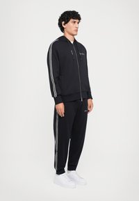 Man standing wearing black tracksuit with white stripes on sleeves and pants, paired with white sneakers, facing right.