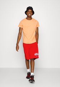T-shirt arancione a maniche corte, pantaloni corti rossi con grafica "USA", cappello a secchiello nero, calze bianche e sneakers neri con accenti rossi.