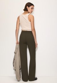 Lys beige ærmeløs top parret med mørke grønne vidde bukser. Personen holder en lysegrå jakke, der fremhæver en flad silhuet og glat tekstur.