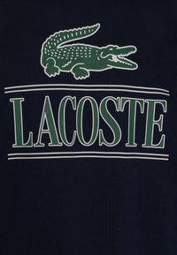 Námořnická modrá látka s zeleným krokodýlem nad textem "LACOSTE" v bílé s zelenými akcenty. Hladký povrch.