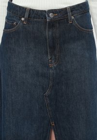 Mørk denimskjørt med frontsplitt, med oransje sømmer, fem lommer og knappelukking i midjen. Glatt tekstur.