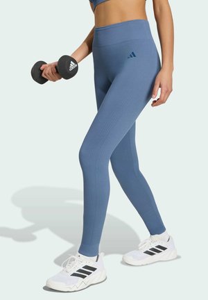 Persona que lleva puestos leggings azules de Adidas y zapatillas blancas de Adidas sostiene una pesa negra de Adidas sobre un fondo claro.