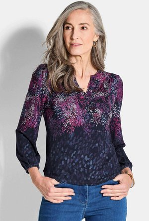Bluse mit floralem Muster in tiefem Marineblau mit lila und blauen Akzenten. Sie hat lange Ärmel, einen V-Ausschnitt und Raffungdetails an den Schultern.