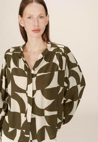 Femme aux cheveux bruns lisses portant une blouse boutonnée ample avec de grandes formes abstraites vert olive et crème, se tenant devant un fond uni.