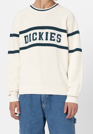 Person trägt einen cremefarbenen Strickpullover mit dunklen horizontalen Streifen und dem Text "DICKIES", kombiniert mit blauen Jeans.