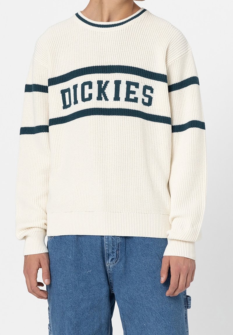 Personne portant un pull en maille crème avec des rayures horizontales foncées et le texte "DICKIES", associé à un jean en denim bleu.
