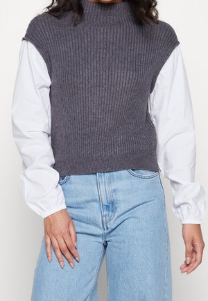 Pull en maille côtelée sans manches gris avec un col haut, superposé à une chemise à manches longues blanche, associé à un jean bleu clair.