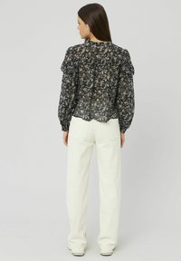 Femme aux longs cheveux bruns portant un blouse noire à motifs floraux avec volants et un pantalon droit taille haute blanc, se tenant de dos.