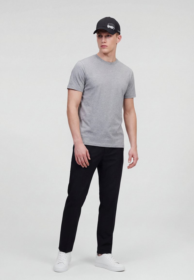 Giovane uomo con maglietta grigia, pantaloni neri, sneakers bianche e cappellino nero in piedi contro uno sfondo chiaro e uniforme.