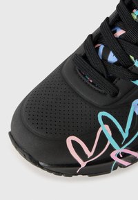 Baskets noires avec un bout perforé, des lacets noirs et des formes de cœurs pastel colorés en rose et bleu sur le côté.
