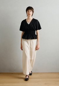 Juoda peplum stilius palaidinė trumpomis rankovėmis ir surinktu liemeniu, derinama su laisvomis kreminėmis kelnių ir juodomis bateliais, ant medinės grindų.