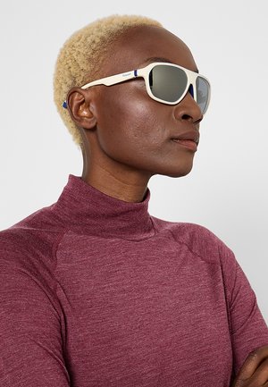 Persona con capelli biondi corti che indossa occhiali da sole con montatura bianca e una maglia bordeaux a collo alto, guarda di lato su uno sfondo semplice.