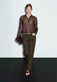 Haut long à manches transparentes marron avec un col et des boutons, associé à un pantalon droit vert olive et des sandales plates. Ceinture marron avec accents métalliques.