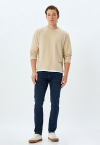 Sudadera beige con mangas largas y cuello redondo; combinada con jeans azul oscuro y zapatillas blancas con detalles en marrón. Diseño simple y limpio.