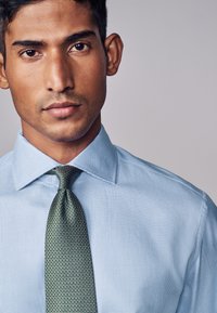Uomo con capelli corti e scuri che indossa una camicia da uomo azzurra con trama e una cravatta verde con motivo, su uno sfondo grigio uniforme.