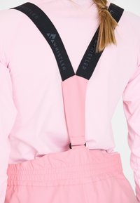 Rosa hängselremmar är fästa vid en matchande klädesplagg, med en svart logotypdetalj. Materialet verkar slätt och elastiskt.