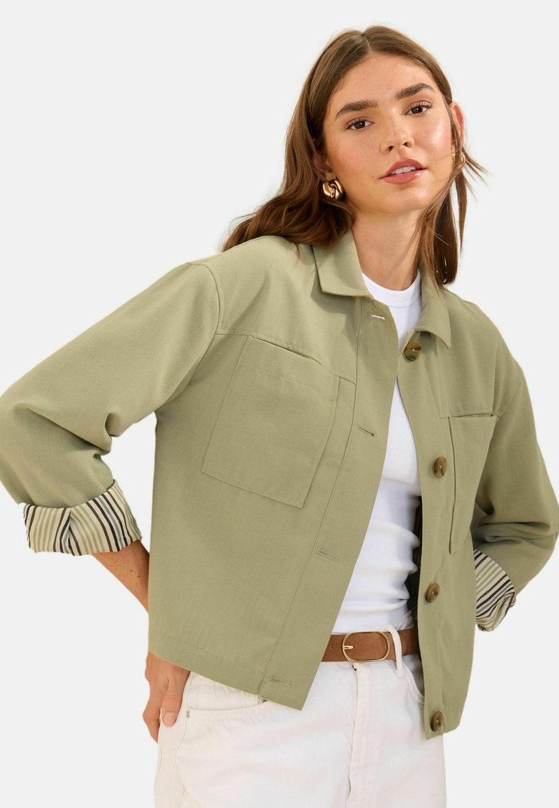 Jeune femme portant une veste boutonnée vert clair sur un haut blanc et un pantalon crème, avec les manches retroussées dévoilant une doublure rayée.