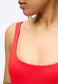 Noor inimene kannab punast Calvin Kleini topi, mille tõttu on nähtav kael, õlg ja osa näost ühtlase tausta ees.