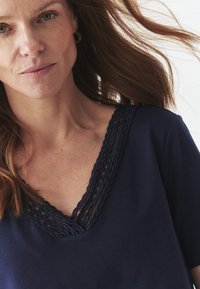 Camiseta de cuello en V azul marino con detalle trenzado en el cuello, mangas cortas y material de algodón suave. Presenta un ajuste relajado y una textura sutil.