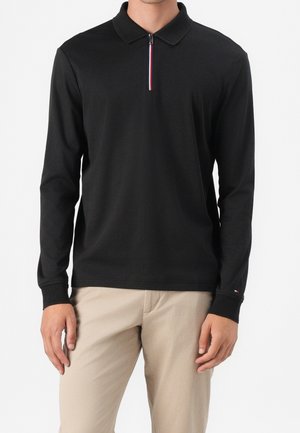 Homme portant un polo noir à manches longues avec une courte fermeture éclair verticale et un pantalon beige, debout sur un fond blanc.