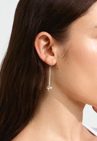Hõbedased ripatsiga kõrvarõngad, mille disain on ahelaga ja lõpeb väikese paelaga. Minimalistlik esteetika poleeritud viimistlusega.