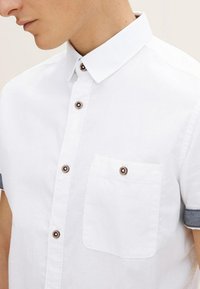 Camisa blanca de manga corta hecha de tejido texturizado, con detalles de botones marrones y un bolsillo en el pecho con ribete gris en las mangas.