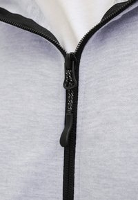 Détail d'une veste zippée grise avec une fermeture éclair noire, une texture de tissu lisse et des accents noirs contrastants le long de la ligne de fermeture.