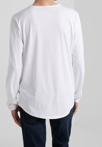 T-shirt à manches longues blanche avec un col rond, texture douce et ourlet arrondi. Accompagné de jeans foncés, présentant une coupe décontractée.