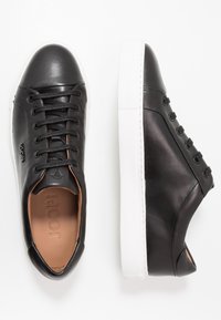 Baskets en cuir noir avec semelles en caoutchouc blanc, dotées de lacets noirs, d'un design minimaliste et d'un logo embossé sur le côté.