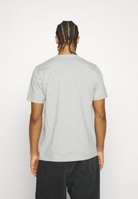Hombre con cabello trenzado, vistiendo una camiseta de manga corta gris claro sencilla y pantalones oscuros, de espaldas contra un fondo blanco.