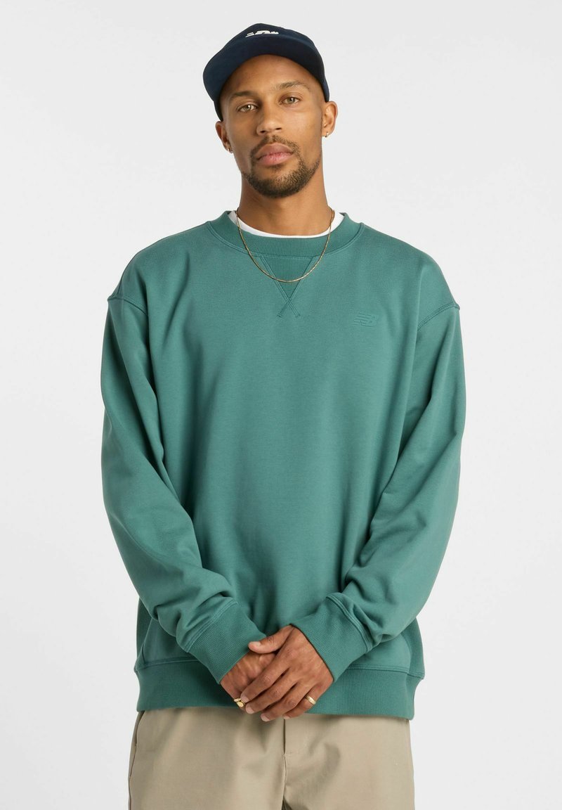 Sweatshirt teal avec un col ras du cou, des poignets et un ourlet côtelés. Présente un logo en relief sur la poitrine. Porté par-dessus un t-shirt blanc.