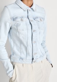 Light blue denim jacket with a fitted design, button closure, two chest pockets, and a soft texture. Collared with adjustable hem.