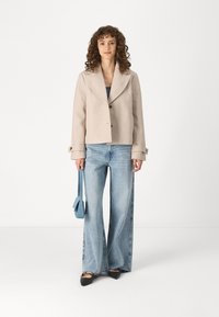 Beige, cropped jacket with a notched collar and three buttons, paired with light blue flared jeans and black flats, holding a blue handbag.