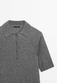 Polo en maille à manches courtes gris avec motif perforé, quatre boutons sur le devant et un col texturé sur fond blanc.