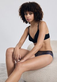 Čierna podprsenka s hlbokým výstrihom v kombinácii s polopriesvitnými tmavomodrými sieťovanými nohavičkami. Model sedí na svetlom štruktúrovanom povrchu, neutrálny pozadí.