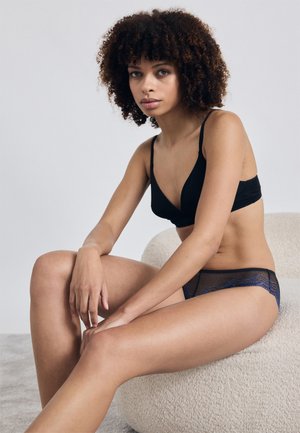 Čierna podprsenka s hlbokým výstrihom v kombinácii s polopriesvitnými tmavomodrými sieťovanými nohavičkami. Model sedí na svetlom štruktúrovanom povrchu, neutrálny pozadí.